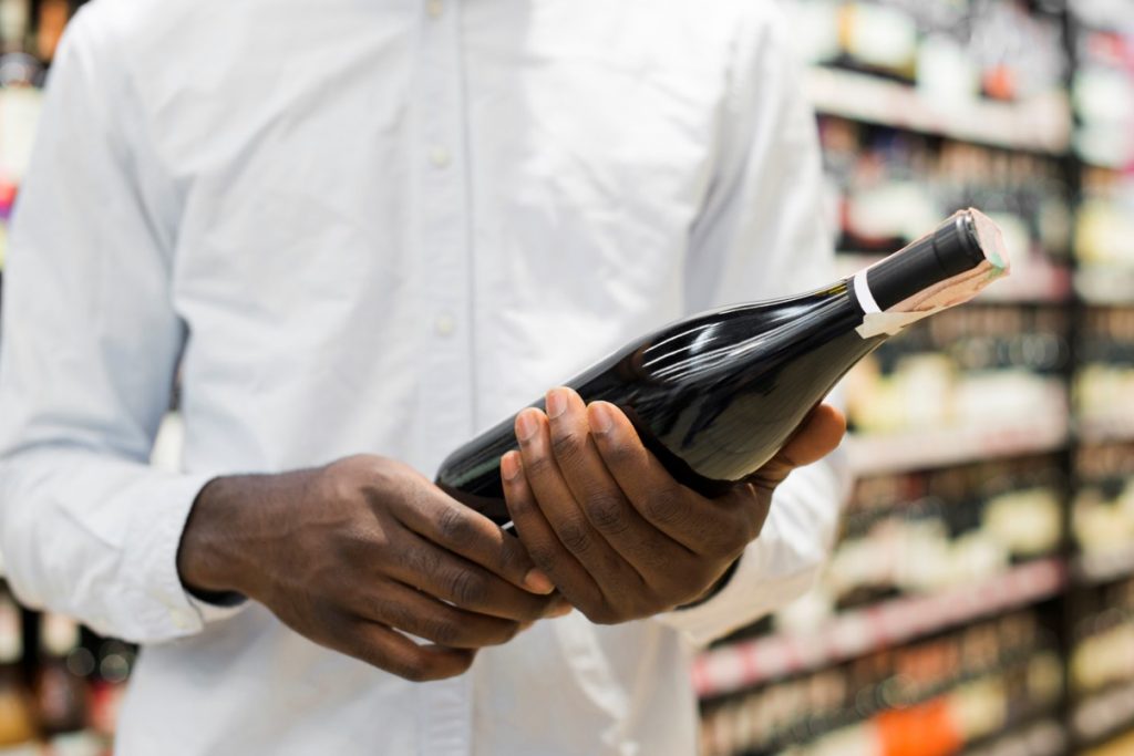 Na imagem vemos um homem segurando uma garrafa de vinho, indicando que ele provavelmente está lendo o rótulo, ao fundo podemos ver que o cenário é um supermercado